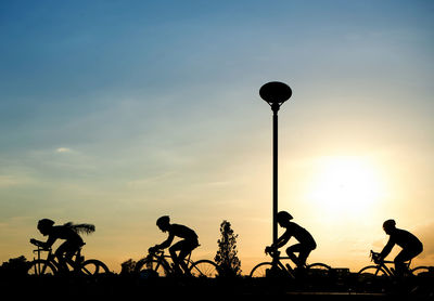 Silhouette people riding bicycles on street against sky during sunset