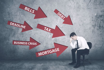 Digital composite image of stressed businessman with red arrow symbols pointing at him against wall