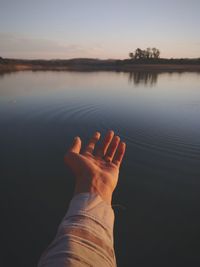 Cropped hand of person gesturing by lake against sky
