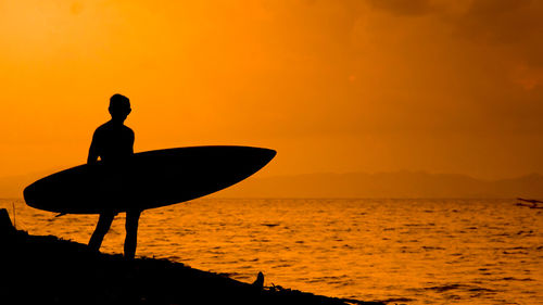 Silhouette man carrying surfboard on shore against orange sky