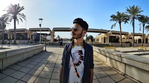 Portrait of young man standing by palm trees against sky