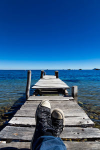 Low section of man standing on retaining wall by sea against clear blue sky