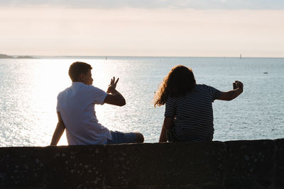 Rear view of friends taking selfie while sitting on retaining wall against sea during sunset