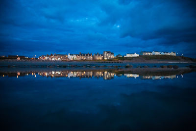 Scenic view of illuminated town and calm lake against cloudy night sky