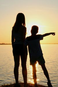 Silhouette boy and girl standing against river during sunset