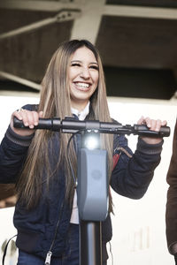 Smiling teenage girl with electric push scooter standing by male friend below bridge