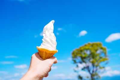Cropped hand of person holding ice cream cone against blue sky