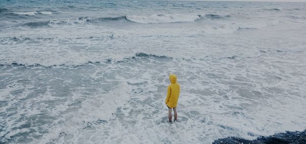 High angle view of woman in yellow raincoat standing on waves at sea shore