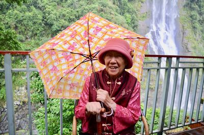 Portrait of woman with umbrella sitting against waterfall