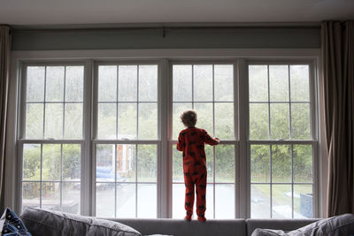 Small boy looks out large living room window at rainy back yard