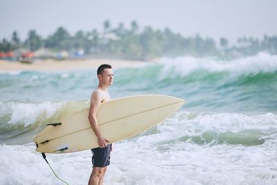 Shirtless man holding surfboard on shore at beach against sea