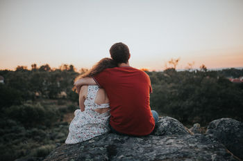 Rear view of couple sitting on rock against clear sky during sunset