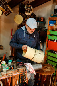 Concentrated male artisan making round wooden shell for drum while working in light professional workshop with abundance of special instruments