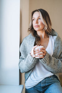 Young woman with blonde long curly hair in cozy grey sweater with cup of tea in hands at home