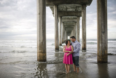 Smiling couple standing on shore under bridge at beach