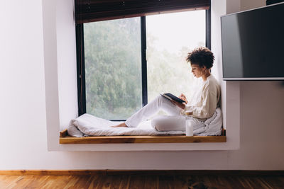 Side view of woman sitting on window, reading and taking notes