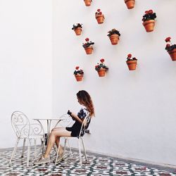 Side view of young woman sitting on chair looking at menu card against potted plants on wall indoors