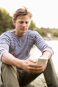 Man text messaging through mobile phone on rock at lake shore