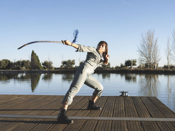 Young woman practicing martial arts with sword against lake at park