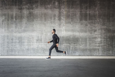 Side view of young man running by gray wall