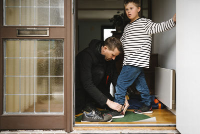 Father helping son with down syndrome to wear shoe standing at doorway