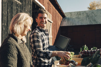 Cheerful male farmer arranging vegetables in crate while standing by friend outside barn