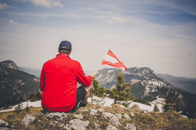 Rear view of man holding flag while sitting on mountain against sky