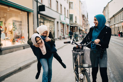 Young woman walking with bicycle looking at cheerful teenage girl giving piggyback to friend on street in city