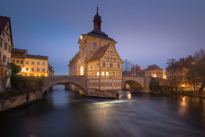 Arch bridge over river amidst illuminated buildings against sky at dusk