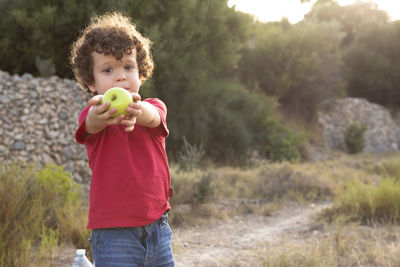 Beautiful curly-haired boy in the field and dressed in red, offering a yellow apple 