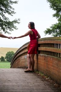 Cropped hand on man reaching girlfriend standing on footbridge at park