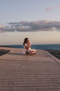 Young woman sitting on shore at sea against sky