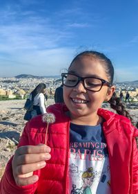 Smiling girl holding dandelion while standing against sky