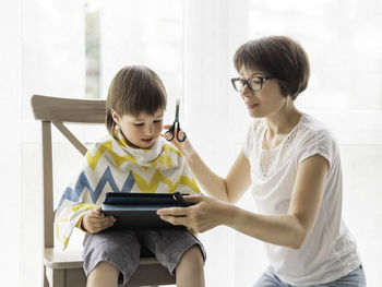 Mother cuts her son's hair by herself. little boy sits with digital tablet. new normal