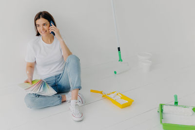 Full length of woman holding color swatch talking on phone while sitting on floor at home