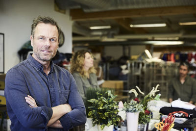 Portrait of confident mature man standing with arms crossed against colleagues working in warehouse