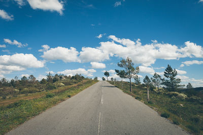 Empty road amidst trees growing on field against sky