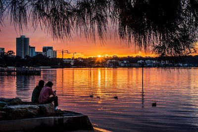 Silhouette people sitting by lake against sky during sunset