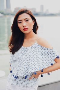 Portrait of young woman standing on footpath against river in city