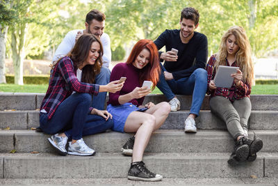 Smiling friends using technologies while sitting on steps