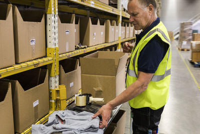 Side view of senior male worker packing merchandise in cardboard box at distribution warehouse