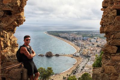 Portrait of man with arms crossed standing by sea against cityscape