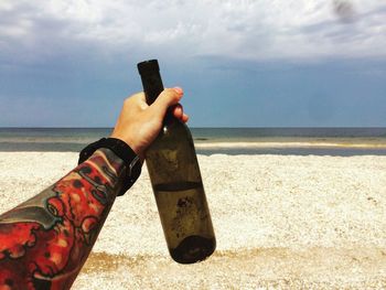 Cropped hand of man with tattoo holding beer bottle at beach