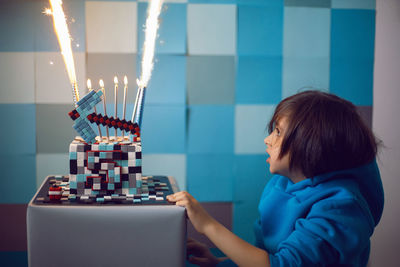 Boy in blue clothes looks at a cake pixels with a pickaxe and candles and lights in the studio.