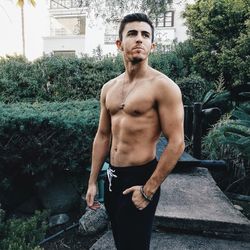 Thoughtful shirtless muscular young man standing against plants