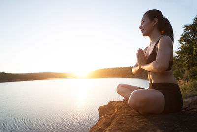 Young woman practicing yoga in prayer position while sitting on rock at lakeshore