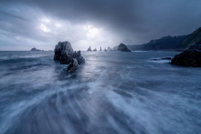 Powerful sea with tough rocky cliffs located amidst slope mountains against cloudy sky in nature of spain