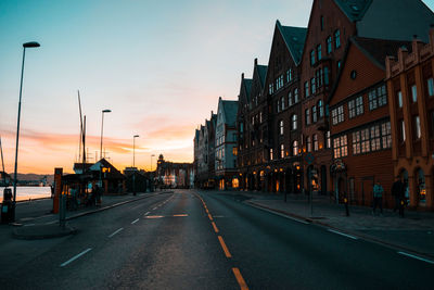 Road by buildings against sky during sunset