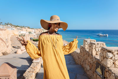 Happy african american woman in yellow dress and sun hat enjoys view of coast of red sea on natural 