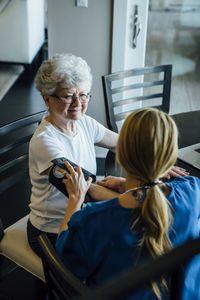 High angle view of home caregiver adjusting blood pressure gauge on senior woman's arm at dining table